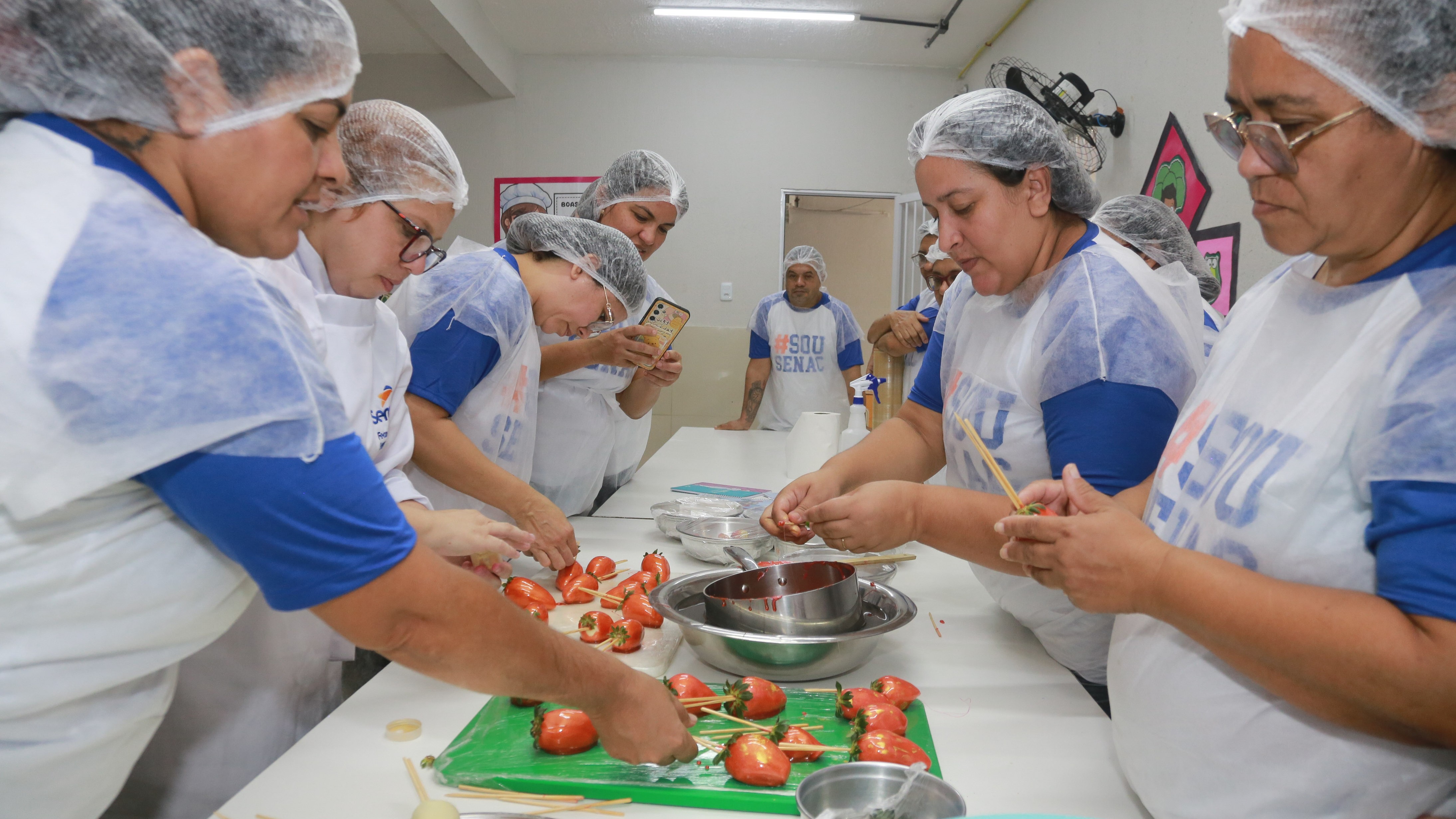 mulheres preparando morango do amor em uma bancada; elas estão molhando o doce num recipiente com a calda vermelha